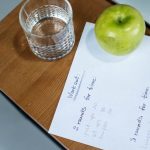 A green apple, glass of water, and workout plan on a wooden desk symbolize healthy living.